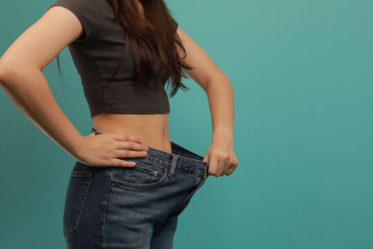 Weight loss transformation concept shown by woman in loose jeans against blue background with confident and happy expression