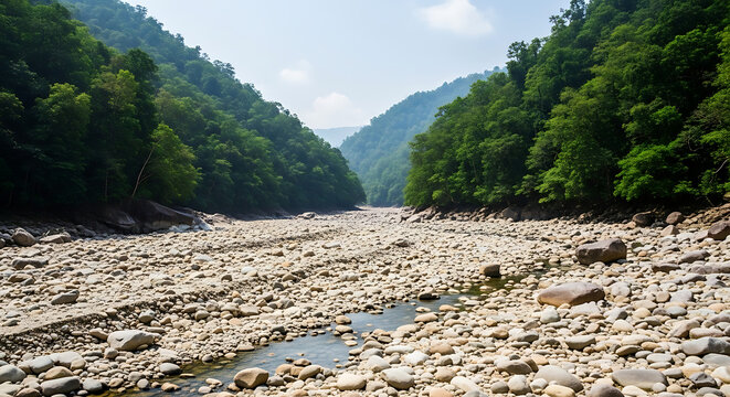 Dry riverbed in mountain valley, rocky river channel with stones and pebbles, green forested hills under blue sky, environmental drought concept