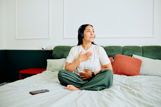 Woman sitting on bed with headphones, hands on chest and stomach, focusing on slow breathing and wellness