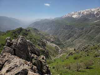 Obraz premium Jagged rocks frame a sweeping spring valley with a nestled mountain village, green slopes, and snow-capped peaks beyond. 📍 Hawraman Takht, Kurdistan, Iran 