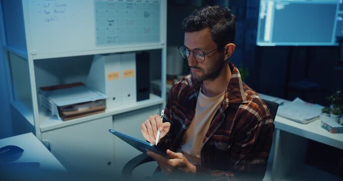 Young Caucasian male programmer use tablet for code design and plan in dark home office at night. AI technology, cybersecurity, software developer, freelance innovation, digital data network concept.