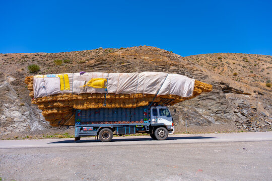 A fully loaded truck carries large hay bales along a road in Morocco. The truck is driving through a rocky area under a clear blue sky in daylight hours.