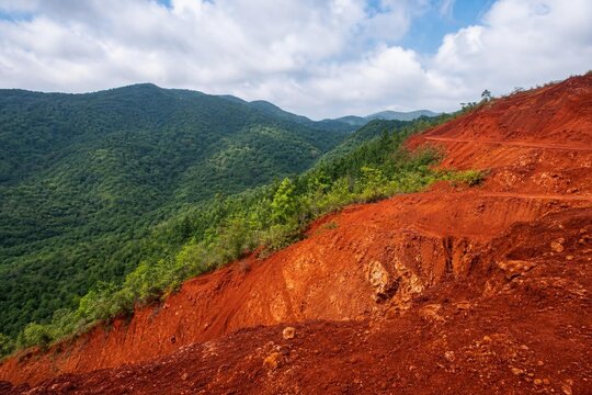 Hillside landscape contrasting rust-red bauxite soil with lush green vegetation, wide view.
