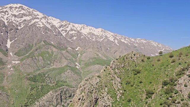 A green rocky ridge faces a massive snow-covered mountain range rising steeply above a deep spring valley under clear blue sky.
📍 Hawraman Takht, Kurdistan, Iran 