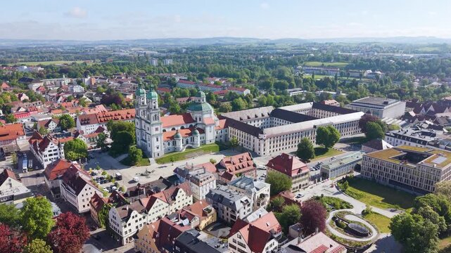 Revealing drone shot of Kempten in Bavaria featuring the Basilica of Saint Lorenz surrounded by historic buildings and a green urban landscape under bright daylight, UHD.