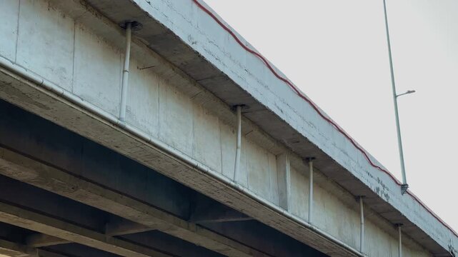 Panning shot showing multiple drainage pipes fixed beneath a reinforced concrete bridge carrying stormwater runoff along the edge of an elevated roadway with urban transportation infrastructure detail