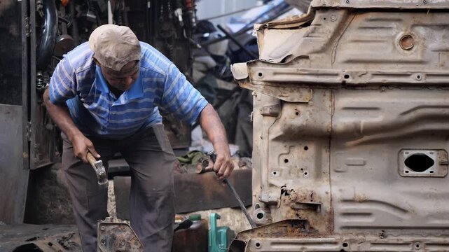  Daylight outdoor shop of an Indian Asian middle aged man or male worker dismantling an old used car or vehicle to recycle the metal scrap at a scrap dealer's workshop in Chor Bazaar, Mumbai, India