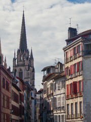 Obraz premium A narrow traditional street in the historic center of Bayonne, France, featuring colorful red shutters and timber-framed houses. The gothic spire of the Bayonne Cathedral rises majestically.