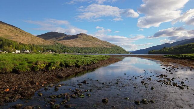 Low and slow drone shot flying down the shallow channel of Loin Water toward the wide expanse of Loch Long and the village of Arrochar on a bright summer evening.