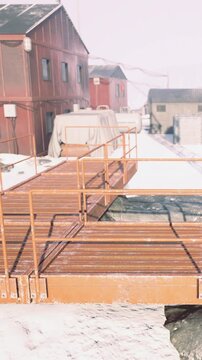 A wooden pathway runs beside buildings in a snowy area. It is cold and the ground is covered in snow. Structures are visible in the background. The scene shows winter conditions.