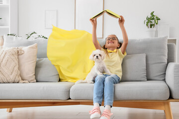 Naklejka premium Cute little happy girl in superhero costume with Maltese dog reading book on sofa at home