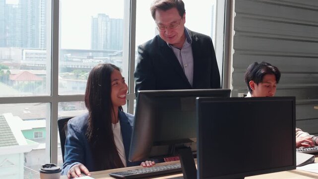 Asian manager behind young businesswoman working at computer, smiling colleagues looking at desktop monitor together, modern office workplace with city, senior boss providing supervision to employee.