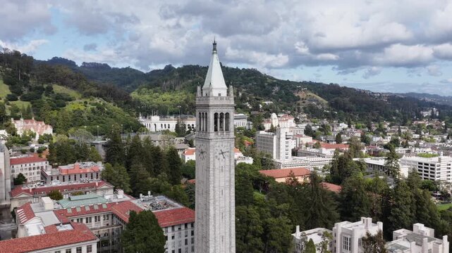 UC Berkeley Campanile clock tower dominating campus with red-tiled buildings, wooded Berkeley Hills and city neighborhoods beyond under partly cloudy skies. California, USA. Aerial orbit shot.