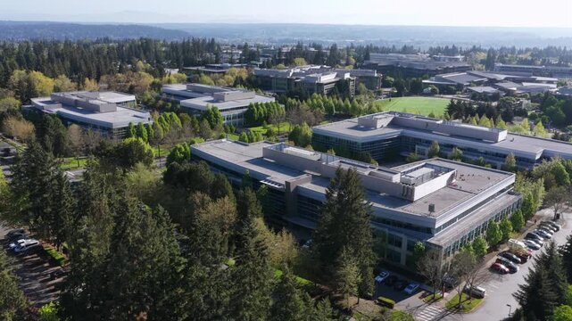 Microsoft headquarters campus with low-rise office buildings spread across tree-lined grounds, green sports field and dense forest to horizon under sunny skies. Washington, USA. Aerial establishing