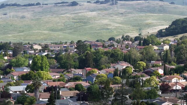 Vallejo residential neighborhood with single-story homes and mature trees, vast green rolling hills rising steeply behind suburban blocks under overcast skies. California, USA. Aerial wide shot.