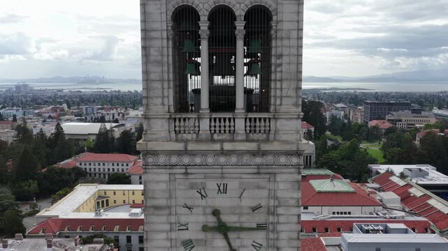 Sather Tower clock face rises above UC Berkeley campus with red-tiled academic buildings and San Francisco Bay glittering beyond under overcast skies, California, USA. Aerial drone rising view.