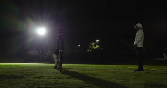 Indian man bowling ball beating batsman, appealing while umpire signaling out, floodlit pitch