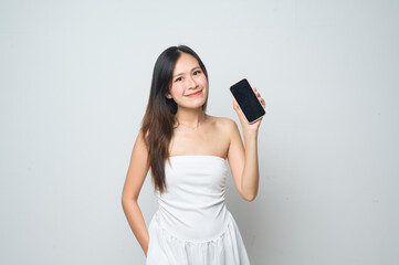 Naklejka premium A young woman in a white dress smiles while holding up a smartphone with a black screen against a plain white background.