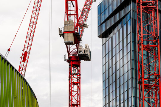 Red construction cranes rise beside a glass facade in an urban development site where architecture and building structure frame the city edge