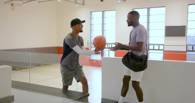 Diverse male friends with basketball and carrying gym bag in gym concourse, with glass railing