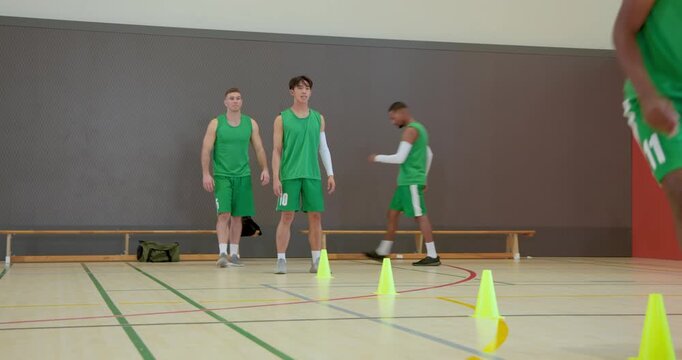 Stepping forward, five male basketball players, dribbling ball through cones in gym, practicing