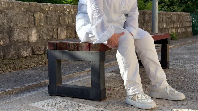 Senior Woman with Knee Pain Sitting on Bench. Senior woman holding a painful knee while sitting on an outdoor bench. Arthritis, joint pain and mobility problem in older age.