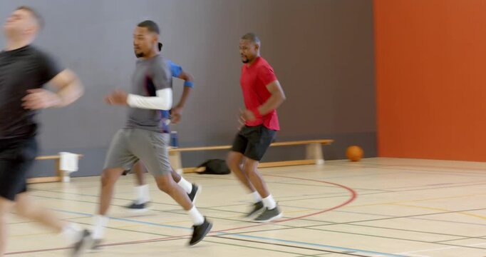Coach signaling five male athletes sprinting gym court past bench holding basketball for agility dri