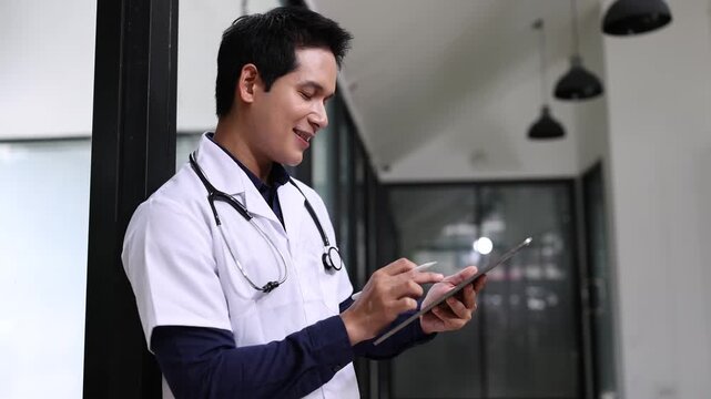 Young Asian male doctor or nurse in blue scrubs using a digital tablet and stylus pen in a modern clinic.