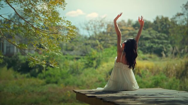 Rear view of a young woman in a white dress sitting on a wooden pier with arms raised in joy, enjoying the sunrise in a lush green garden. Concept of freedom, wellness, and mental health in a natural