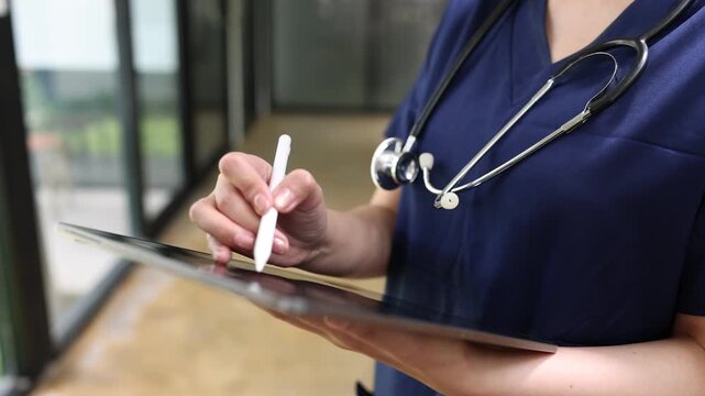 Young Asian female doctor or nurse in blue scrubs using a digital tablet and stylus pen in a modern clinic.