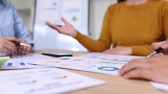 A group of young Asian business colleagues working together around a table in a modern office, analyzing financial charts, taking notes, and discussing data with bright, positive expressions.