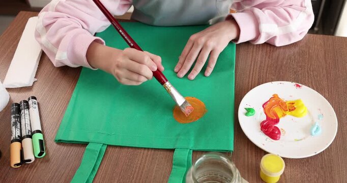 Girl painting on green reusable eco tote bag at home. Creative young artist upcycling fabric shopper with cute custom animal food illustration. Teenager practicing sustainable zero waste lifestyle