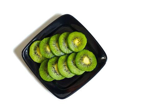 Top view of fresh sliced kiwi (Actinidia deliciosa) arranged on a black square plate, showcasing vibrant green flesh, tiny seeds, and neat presentation isolated on white background with copy space