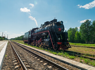 Naklejka premium Old black steam locomotive moving along parallel train tracks next to a dense green forest