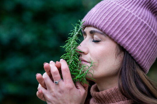 Portrait of beautiful woman wearing pink knit hat smelling fresh rosemary with closed eyes