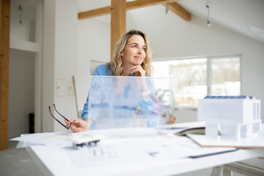 Thoughtful architect with hand on chin and transparent tablet PC leaning at desk