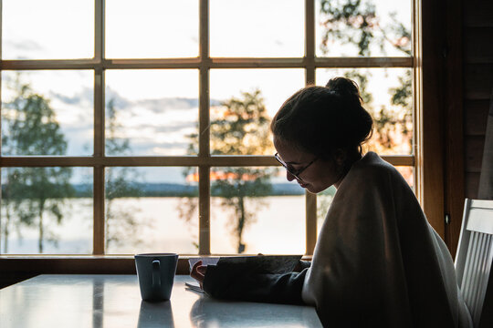 Finland, Lapland, young woman sitting at the window at a lake looking in diary