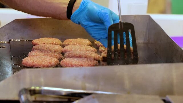 Chef using spatula to flip raw meat patties on professional flat top metal griddle in commercial kitchen.