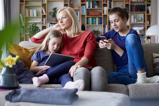 Sad mother sitting on couch with her daughters, playing with mobile devices
