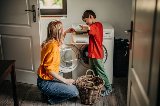 Son pouring detergent in washing machine with mother crouching at home