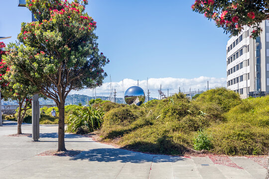 The KIMI / You Are Here reflective sphere sculpture sits among greenery and flowering trees beside modern buildings and a marina in Wellington, New Zealand