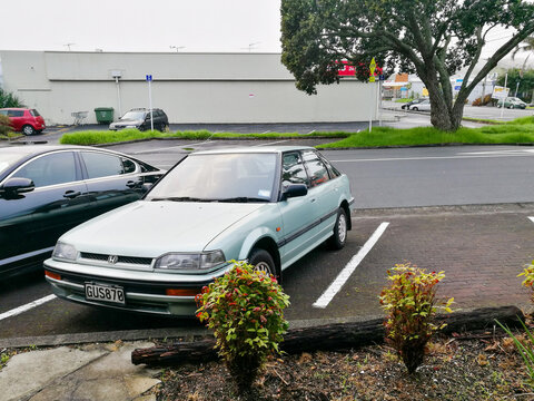 A light green Honda sedan sits in a parking space in Auckland, New Zealand, surrounded by other vehicles and urban greenery