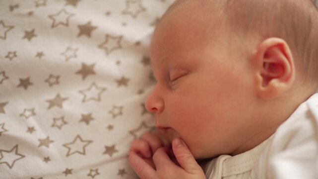 Newborn baby sleeping peacefully in a cradle, eyes closed and mouth slightly open, hands near face, wearing a white bodysuit on star patterned sheets, cozy and calm