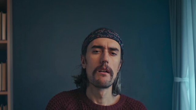 Long-haired man with mustache horseshore in patterned headband and burgundy knit sweater, sitting indoors by blurred bookshelves, looking skeptically at camera in natural daylight portrait