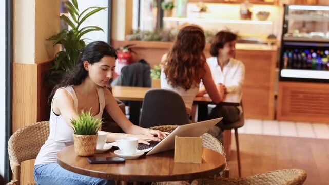 Young woman is sitting in a coffee shop with a laptop and is carefully typing. European woman works remotely from a cafe. Woman communicates on the Internet in social networks