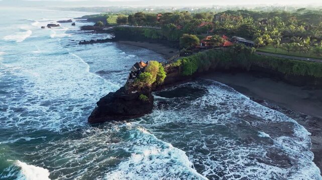 Aerial view of sea temple perched on volcanic rock formation above natural sea arch surrounded by crashing ocean waves in early morning light along the Bali coastline.
