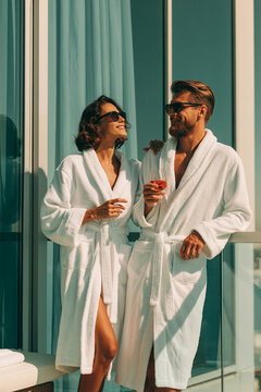 Luxury couple in white robes, hotel balcony, wine glasses, summer vacation lifestyle, elegant resort