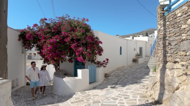 A diverse couple of a man and a woman explores the picturesque alleys of Lefkes Paros, Greece, as vibrant flowers bloom under the bright sun. Discover the beauty of the Cyclades 