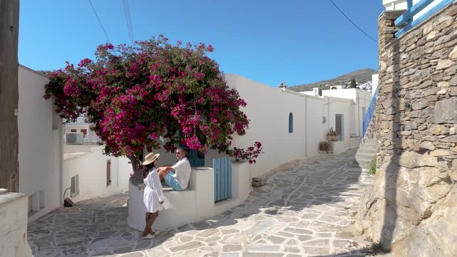 A diverse couple discovers the enchanting alleyways of Lefkes Paros, Greece, as a traveler admires vibrant bougainvillea. This picturesque setting offers a glimpse into the beauty of Cyclades islands