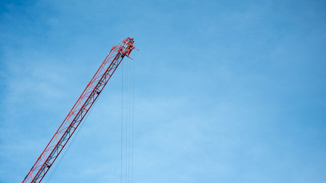 Red lattice boom construction crane jib against a clear blue sky, showing steel structure, pulleys, and cables. Industrial engineering, building, and power.